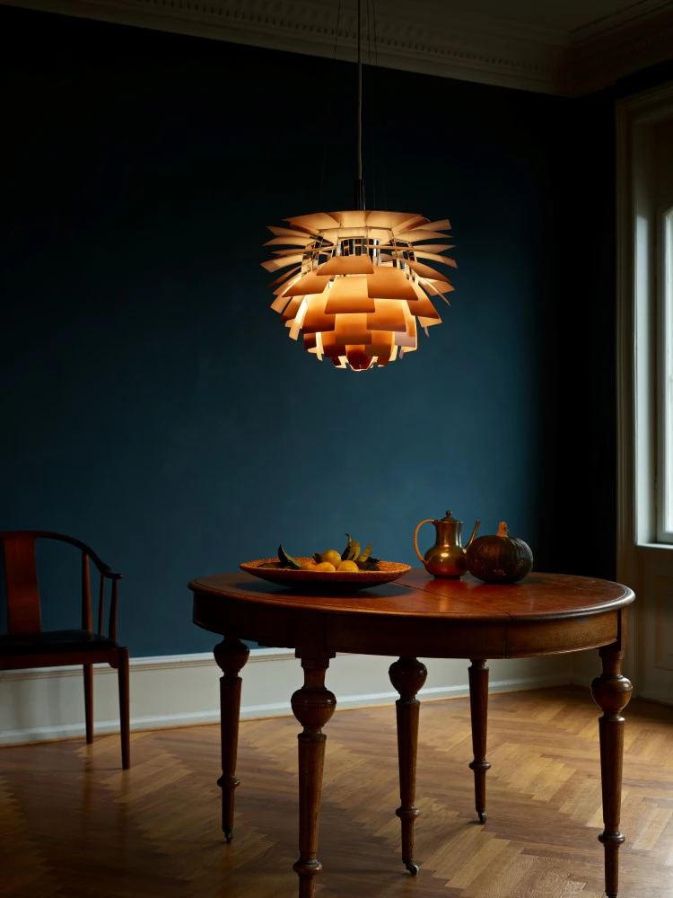 A copper-toned PH Artichoke pendant glows above a round wooden table in a dark dining room. © 2026 Louis Poulsen.