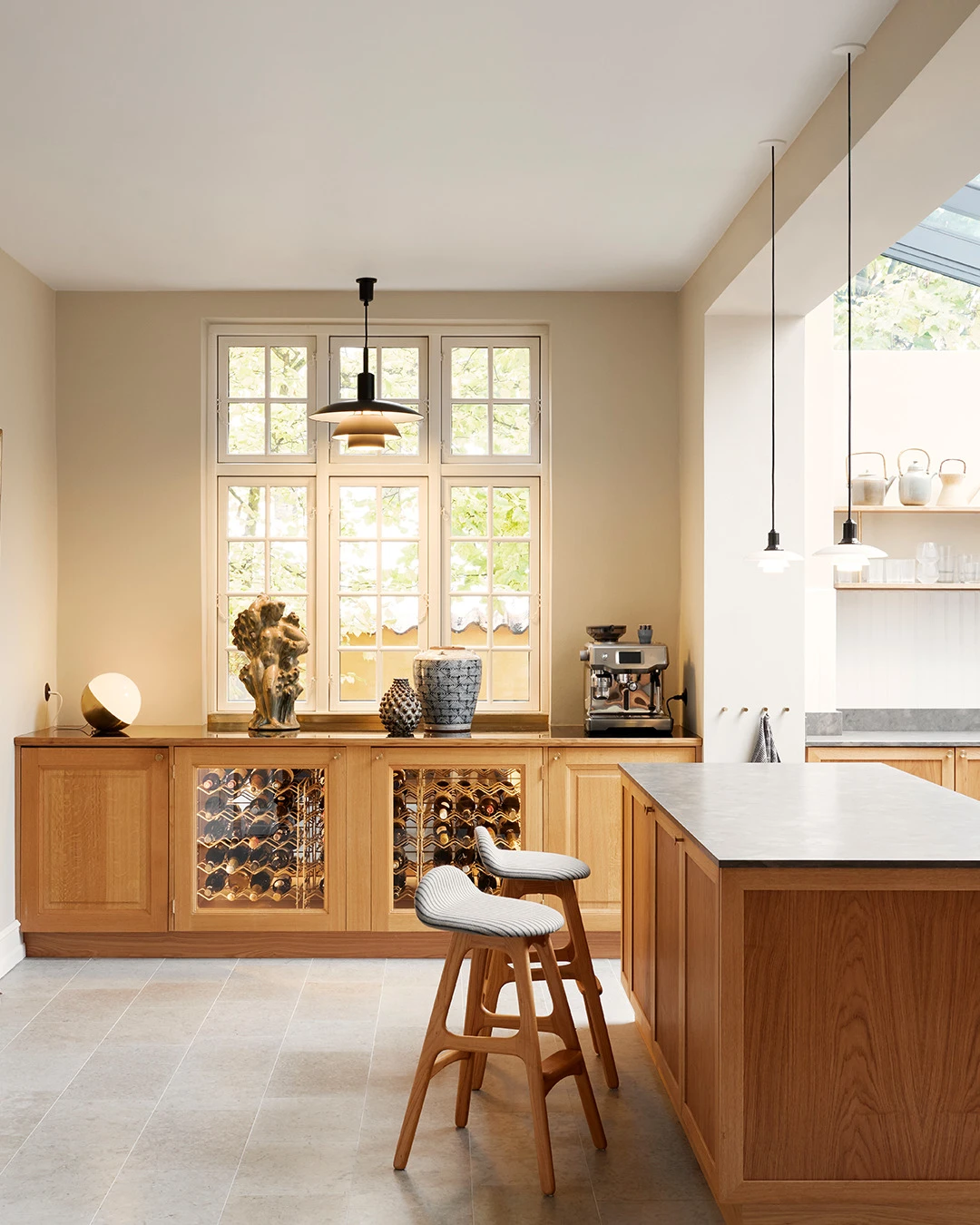 A black PH 5/5 Pendant and two white PH 5/5 Pendants hang in a bright kitchen with wooden cabinets and an island. © 2026 Louis Poulsen.