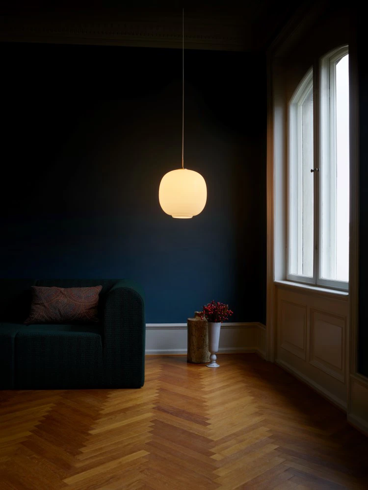 An opal-white Moser Pendant glows above a dark seating area with herringbone flooring and tall windows. © 2026 Louis Poulsen.