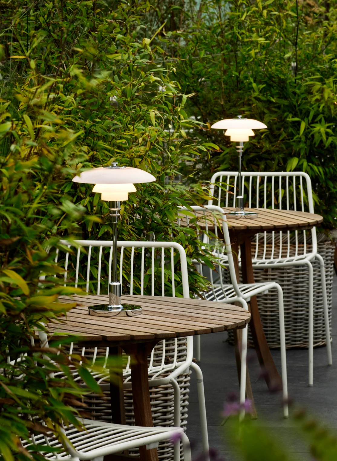 Twee draagbare PH 2/1 lampen verlichten houten tafels tussen tuinmeubilair en dicht groen. © 2026 Louis Poulsen.