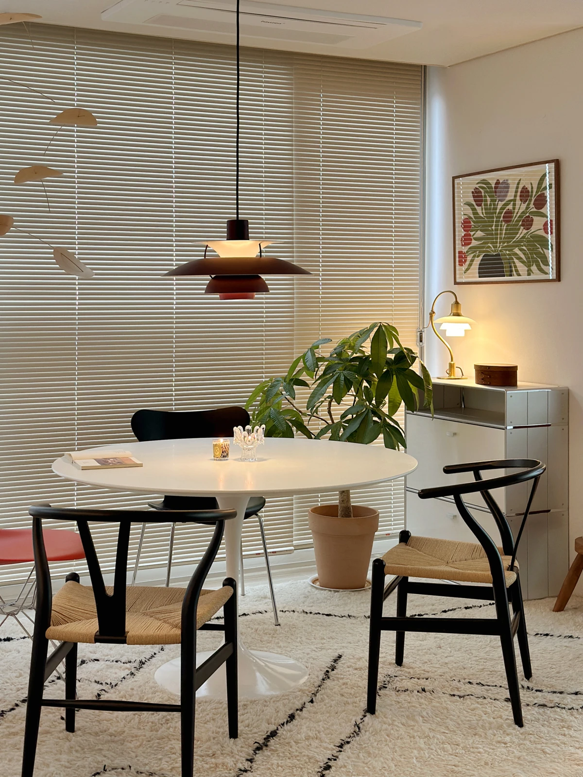 A dark PH 5 pendant hangs above a round dining table in a bright room with wooden chairs and leafy plants. © 2026 Louis Poulsen.