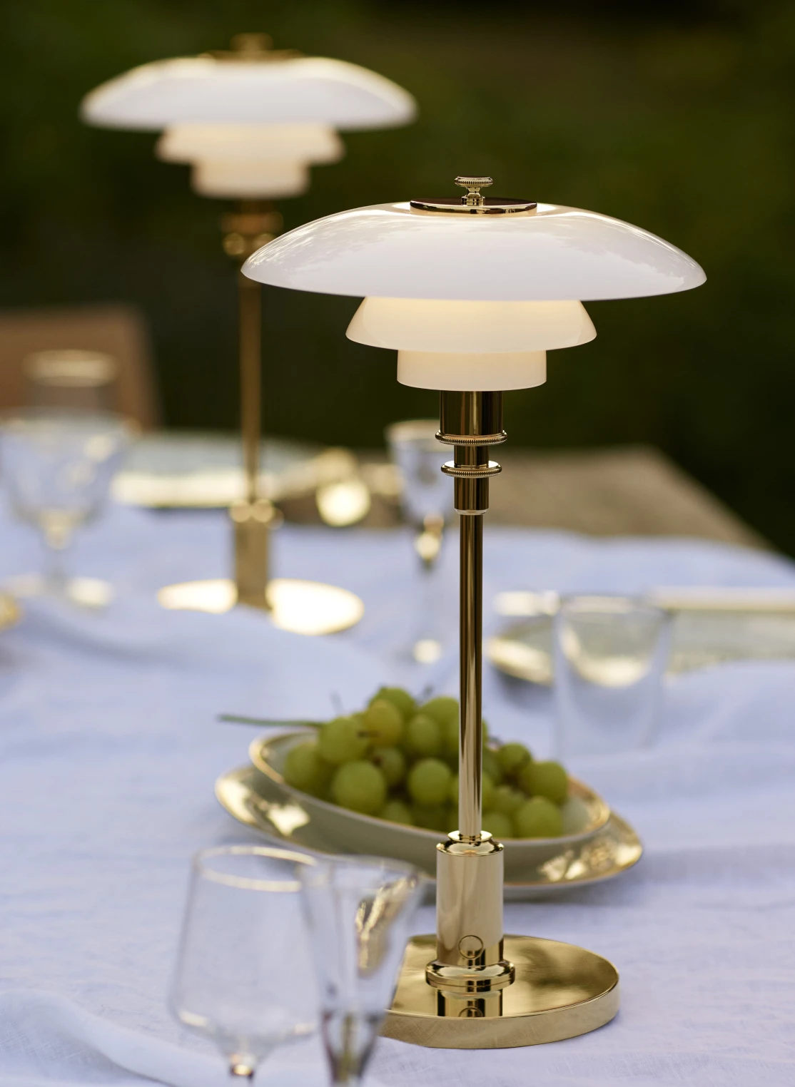 PH 2/1 Portable Lamp i mässing med vita glasskärmar lyser upp ett dukat matbord med en skål vindruvor. © 2026 Louis Poulsen.