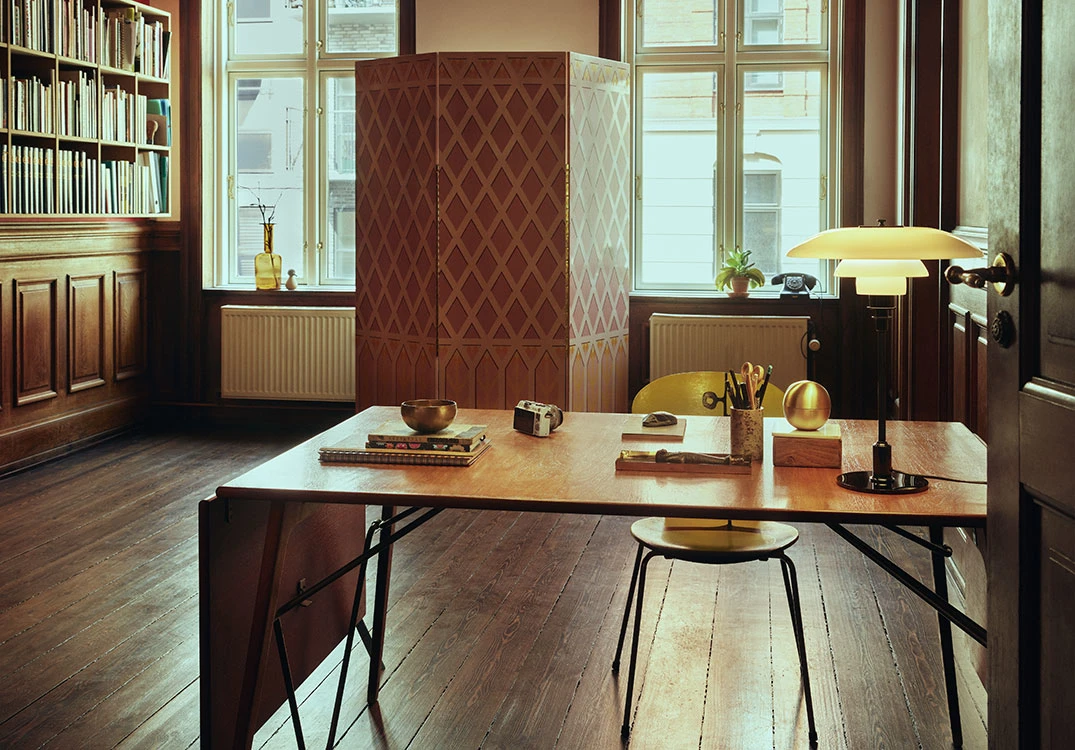 Eine PH 3½-2½ Tischleuchte in Schwarz und Messing steht auf einem Schreibtisch in einem klassischen Arbeitszimmer. © 2026 Louis Poulsen.
