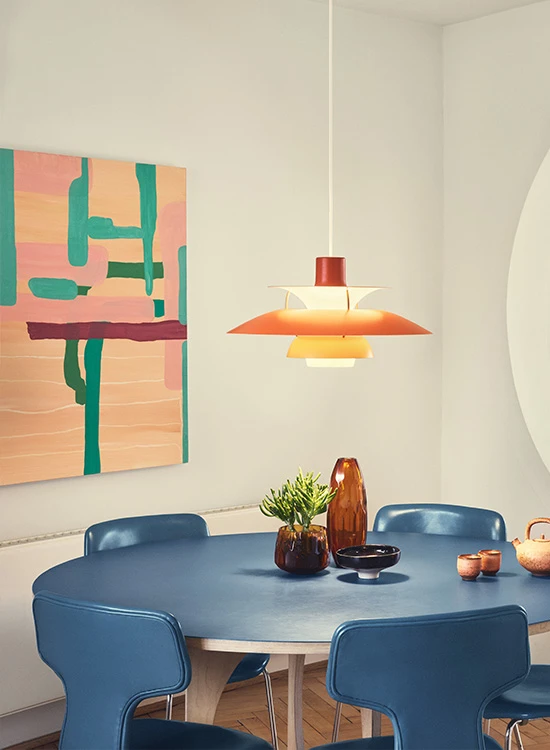 Eine orange PH 5 Pendelleuchte erhellt einen blauen Esstisch mit passenden Stühlen und einem farbenfrohen Bild an der Wand. © 2026 Louis Poulsen.