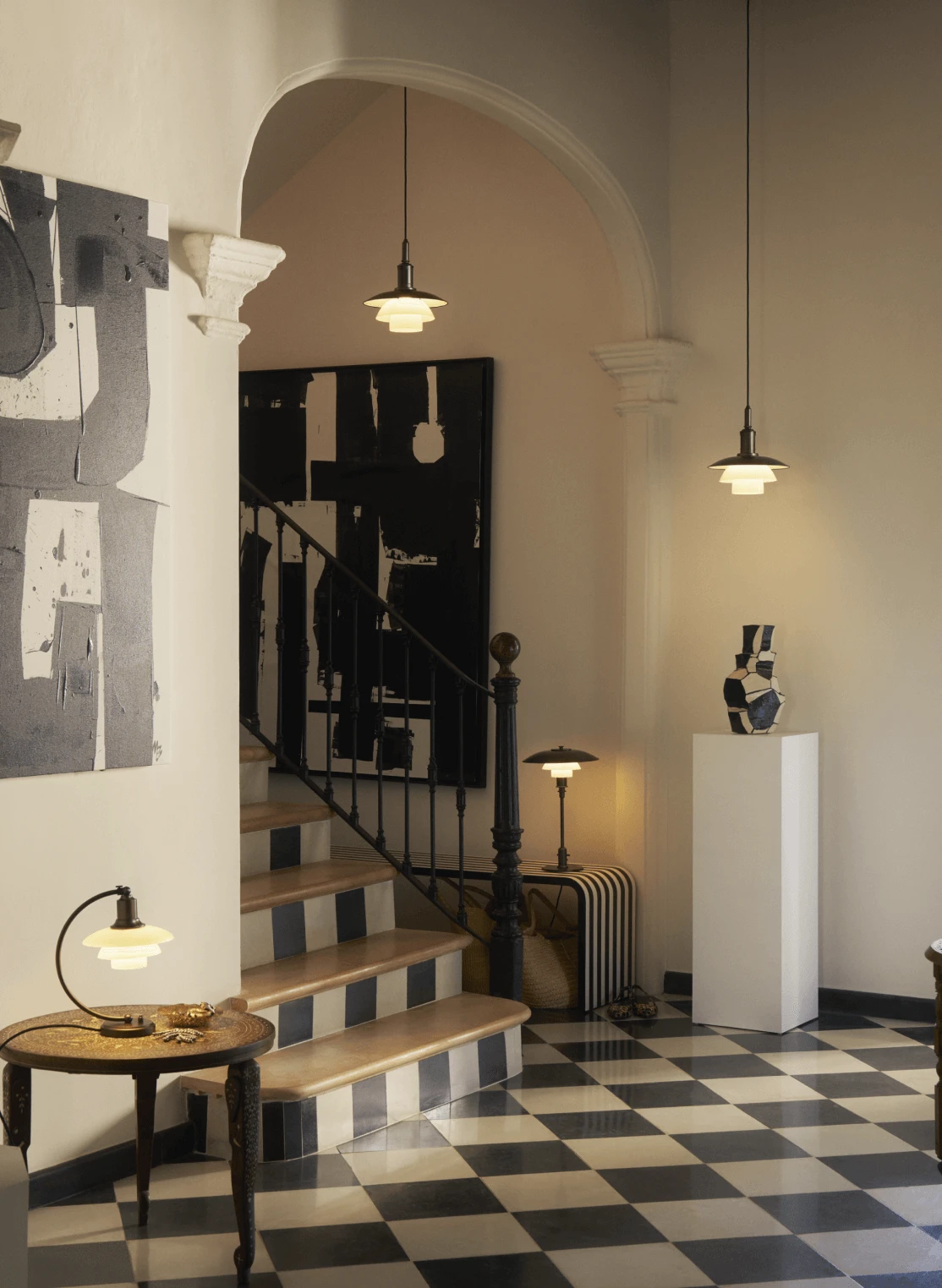 Softly glowing PH Aged brass lamps in a checker-floored interior with art and staircase, © 2025 Louis Poulsen.