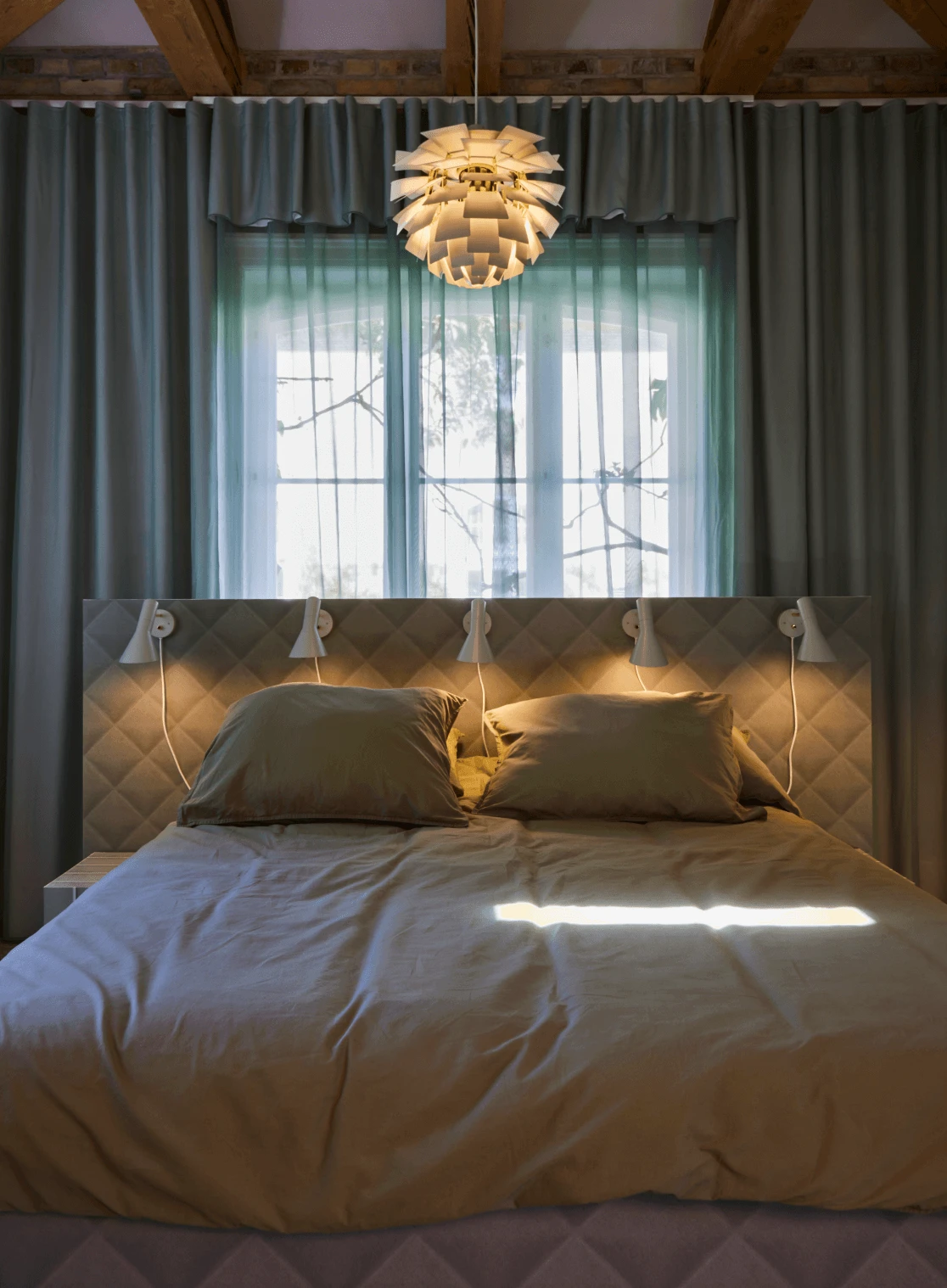 A warm-lit PH Artichoke pendant hangs above a bed with a quilted headboard and wall lights. © 2026 Louis Poulsen.