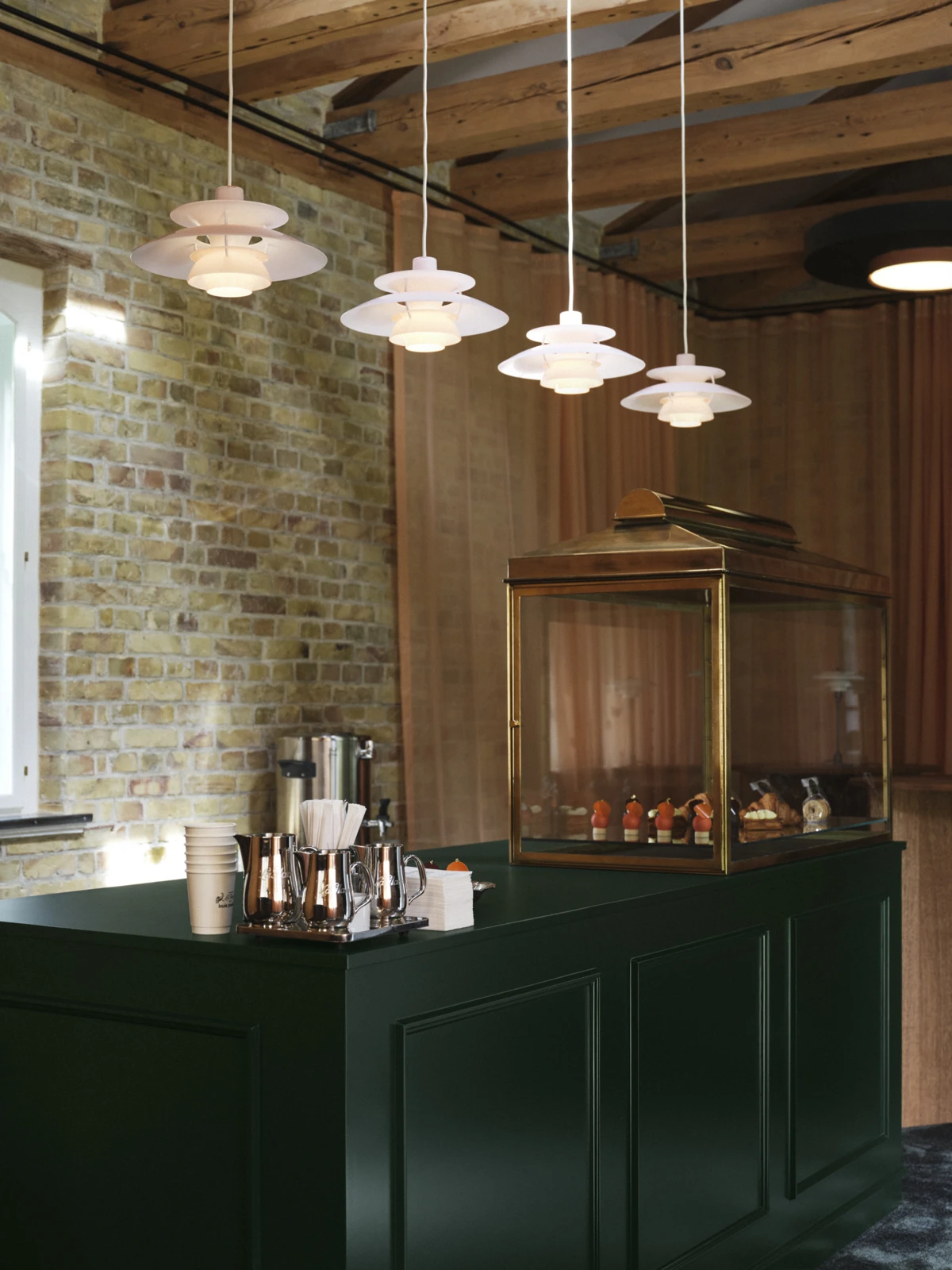 Four white PH 5 pendants hang above a dark green café counter beneath exposed timber beams and beside a brick wall. © 2026 Louis Poulsen.