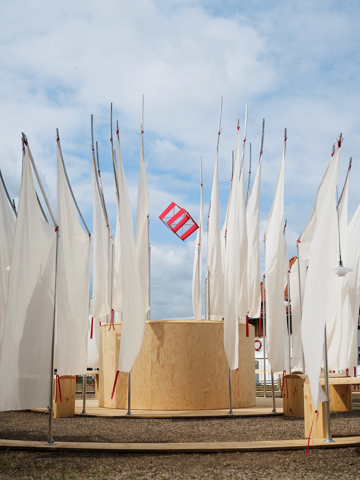 A white Above Pendant hangs among tall white sails around a wooden pavilion beneath a blue sky. © 2026 Louis Poulsen.