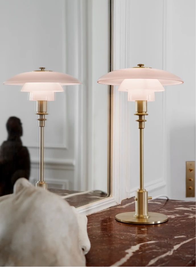A PH 3/2 Pale Rose Table Lamp with a brass stem sits on a dark red marble surface beside a mirror. © 2026 Louis Poulsen.
