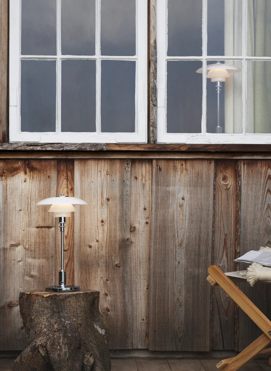 Een PH 2/1 Portable Lamp van opaalglas en chroom brandt tegen een rustieke houten wand. © 2026 Louis Poulsen.