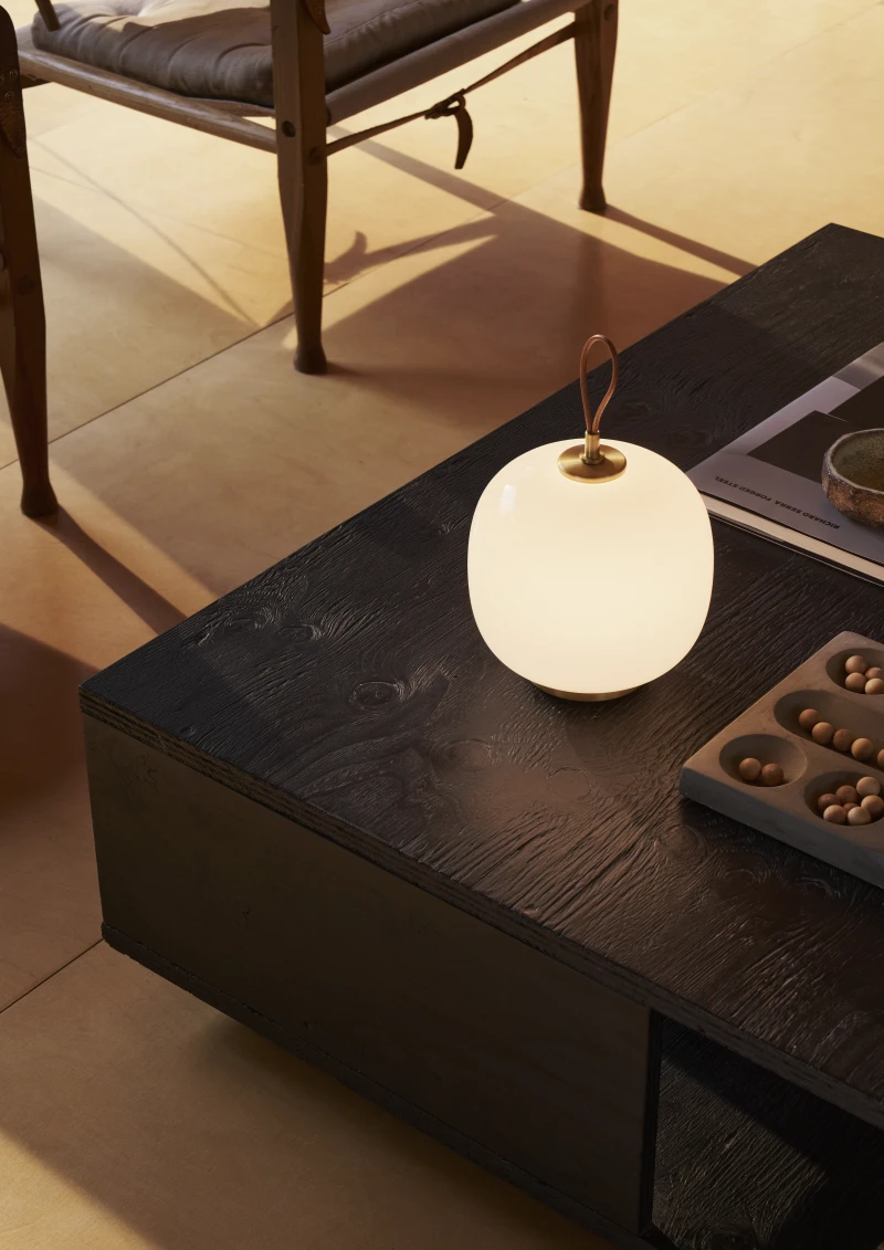 Een VL 45 Radiohus draagbare lamp met een opaalwitte glazen bol en messingdetails geeft licht op een donkere houten tafel. © 2026 Louis Poulsen.