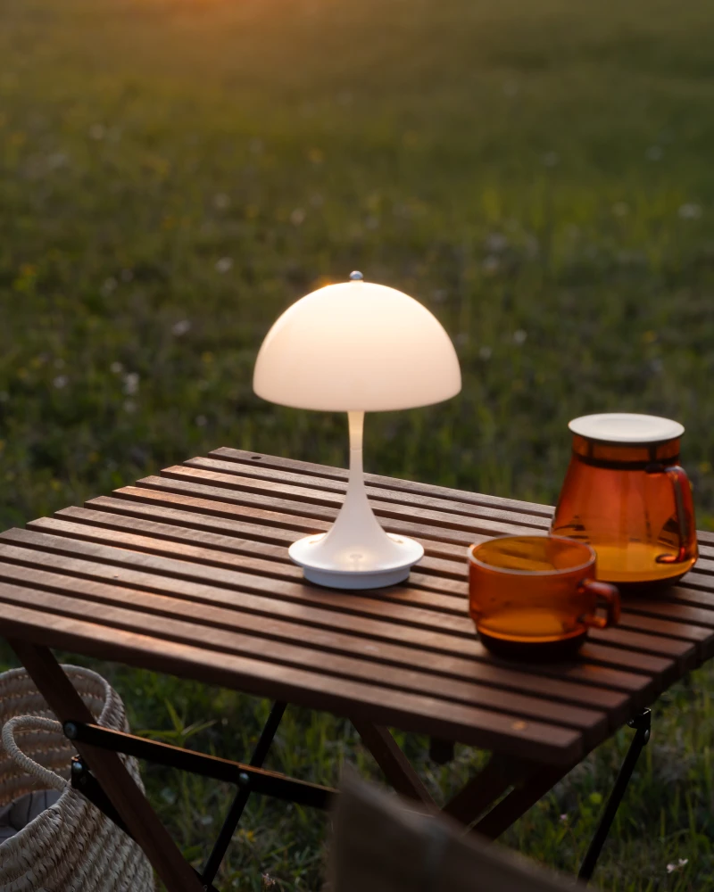 Een witte Panthella 250 draagbare lamp verlicht een houten tuintafel naast amberkleurig glaswerk. © 2026 Louis Poulsen.