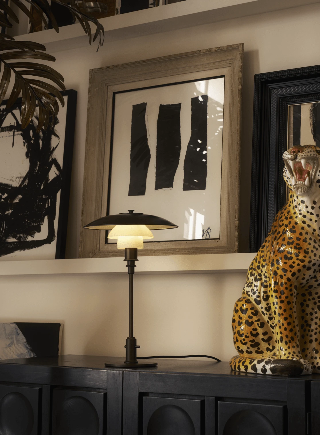 Table lamp on dark cabinet beside bold artwork and ceramic leopard, © 2025 Louis Poulsen.
