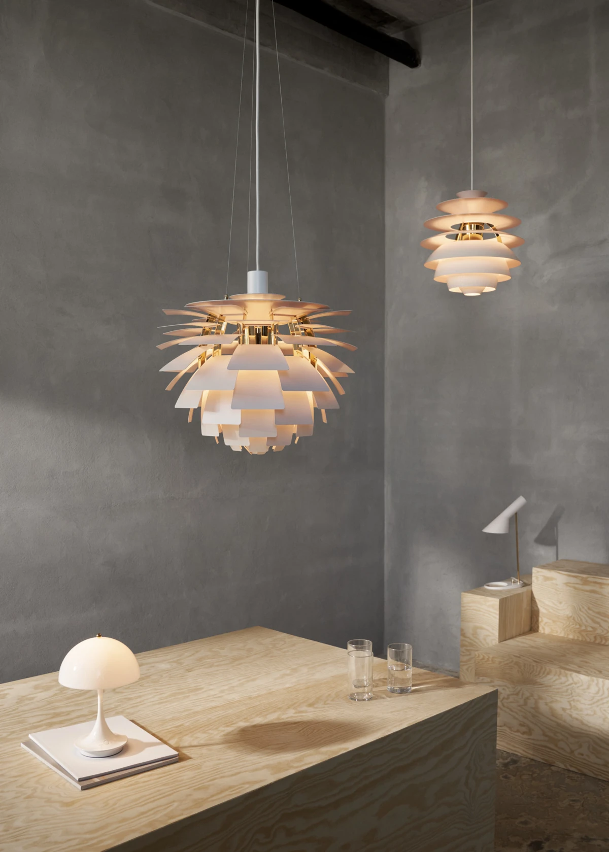 En PH Artichoke-pendel og en PH Snowball-pendel lyser over træpodier med en AJ Mini Anniversary Edition-bordlampe og en Panthella 160 Portable Lamp Anniversary Edition. © 2026 Louis Poulsen.