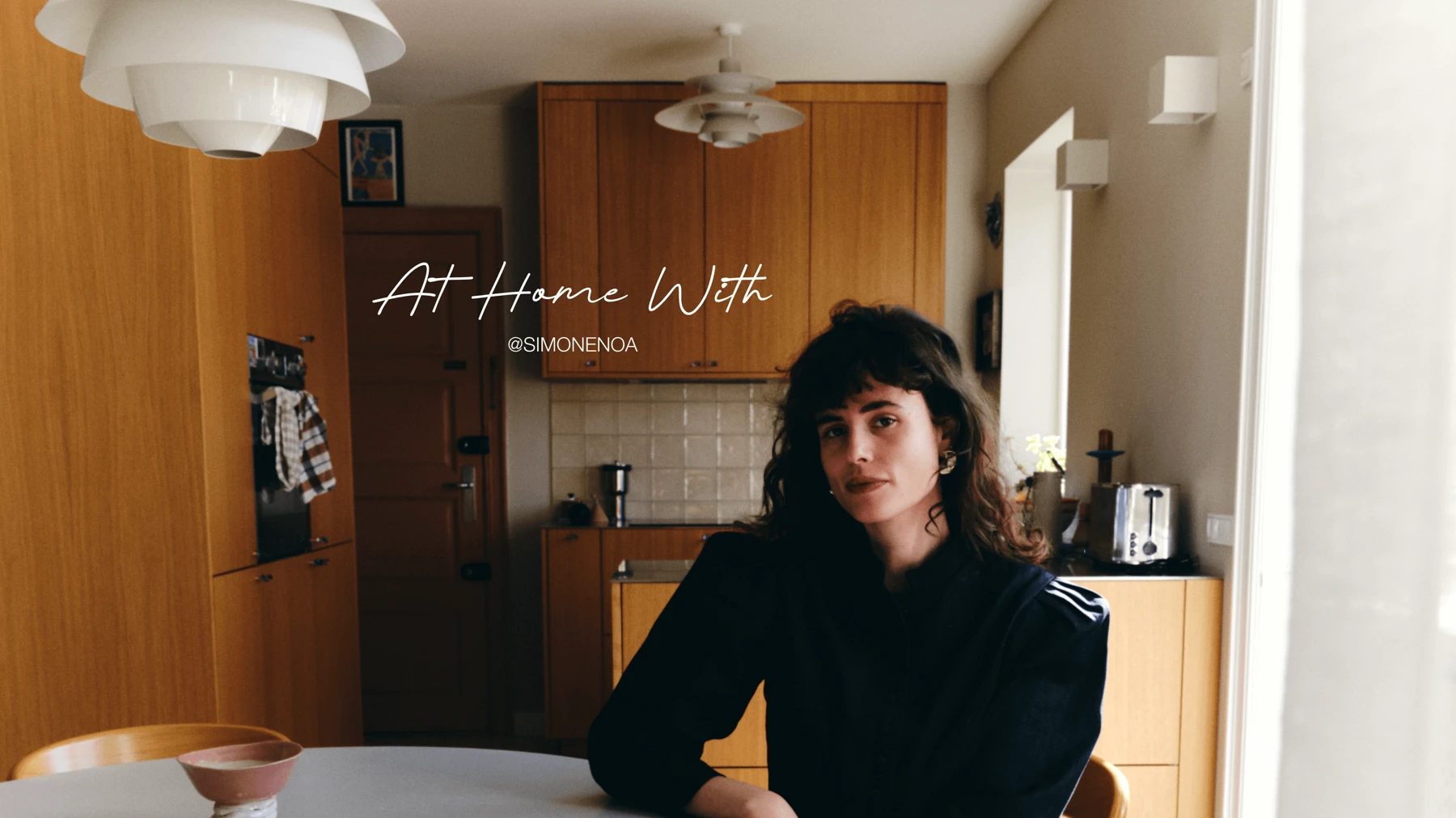 Two white PH 5 pendants hang in a wood-fronted kitchen, where Simone Noa sits at the table. © 2026 Louis Poulsen.