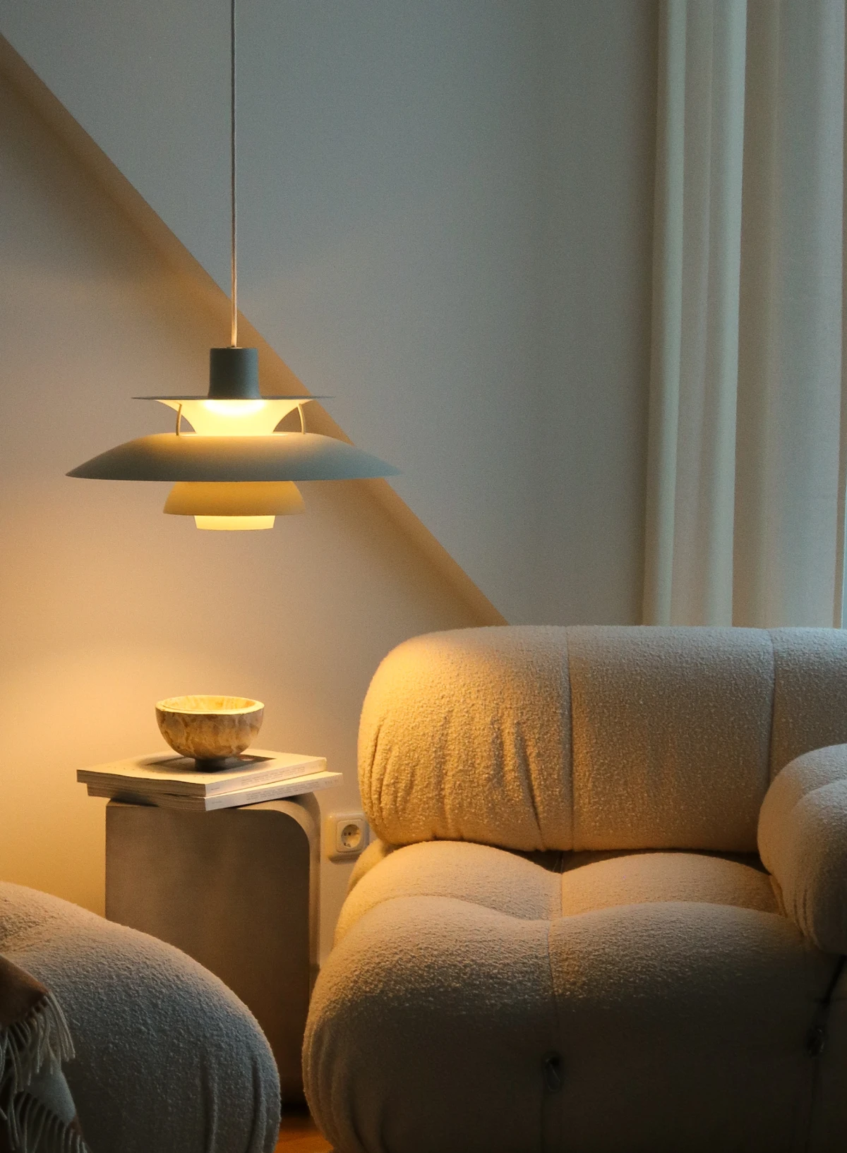 A grey PH 5 pendant casts warm light over a small side table in a cosy living room with soft lounge chairs. © 2026 Louis Poulsen.