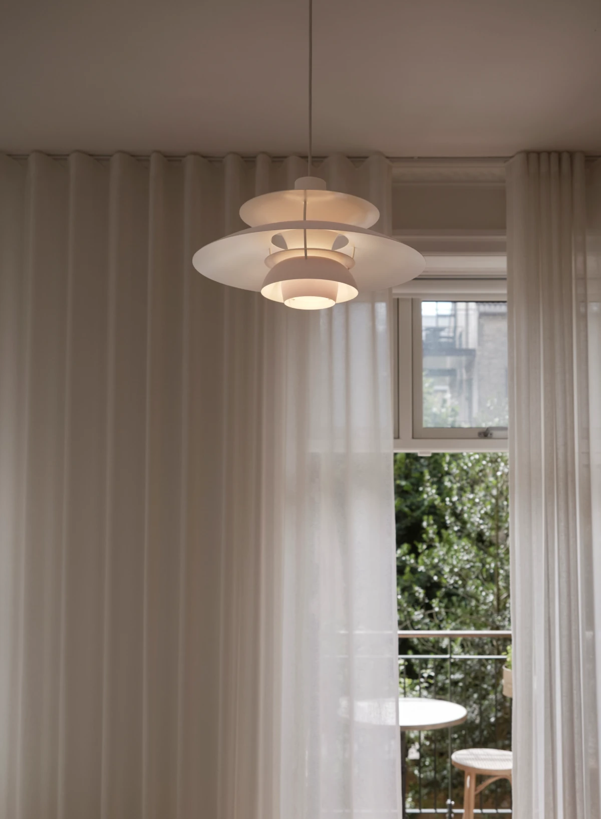 A white PH 5 pendant casts a warm glow in front of sheer curtains and a window overlooking greenery. © 2026 Louis Poulsen.