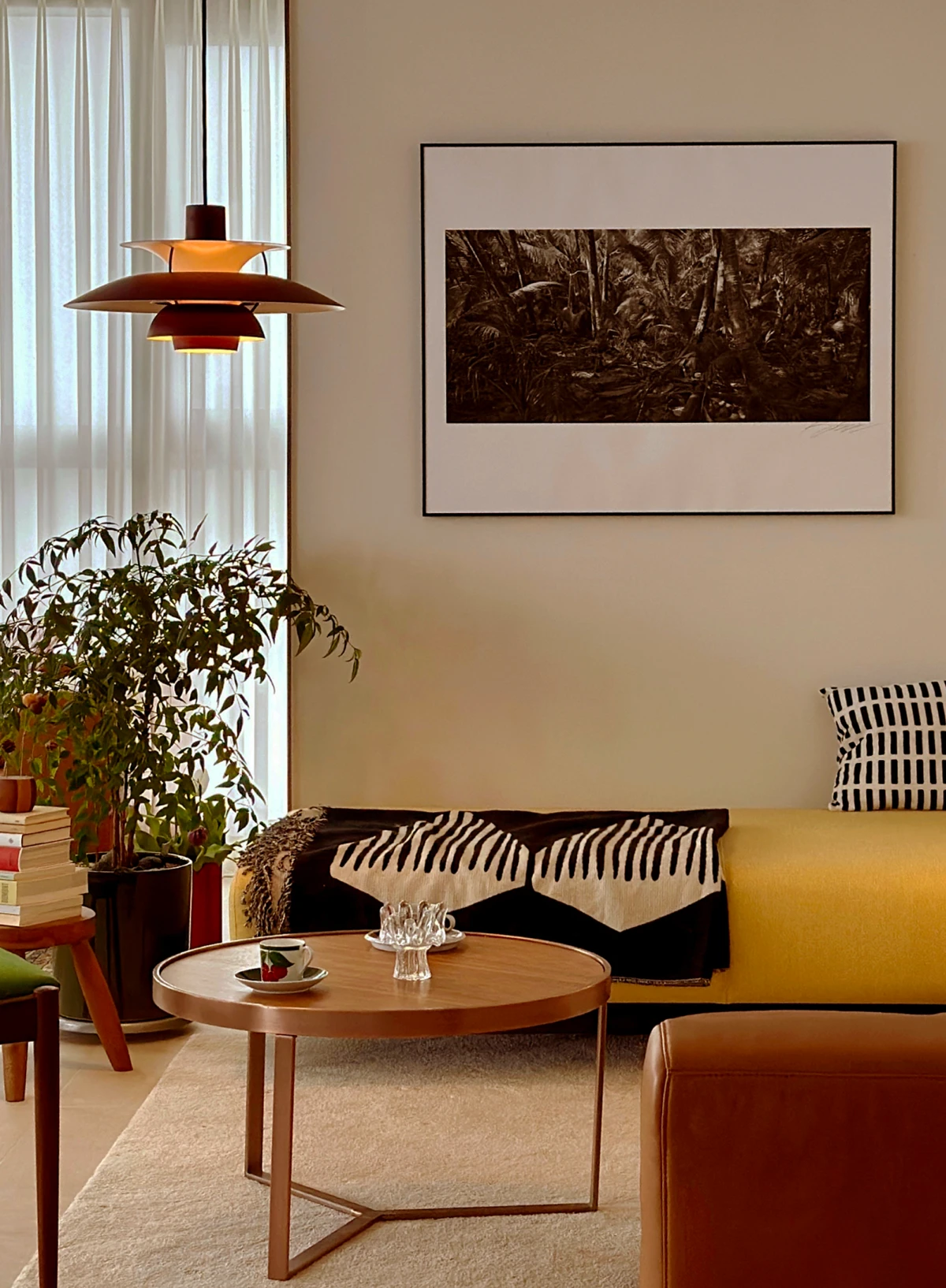 A PH 5 pendant in warm red and orange tones hangs by the window in a living room with plants and a yellow sofa. © 2026 Louis Poulsen.