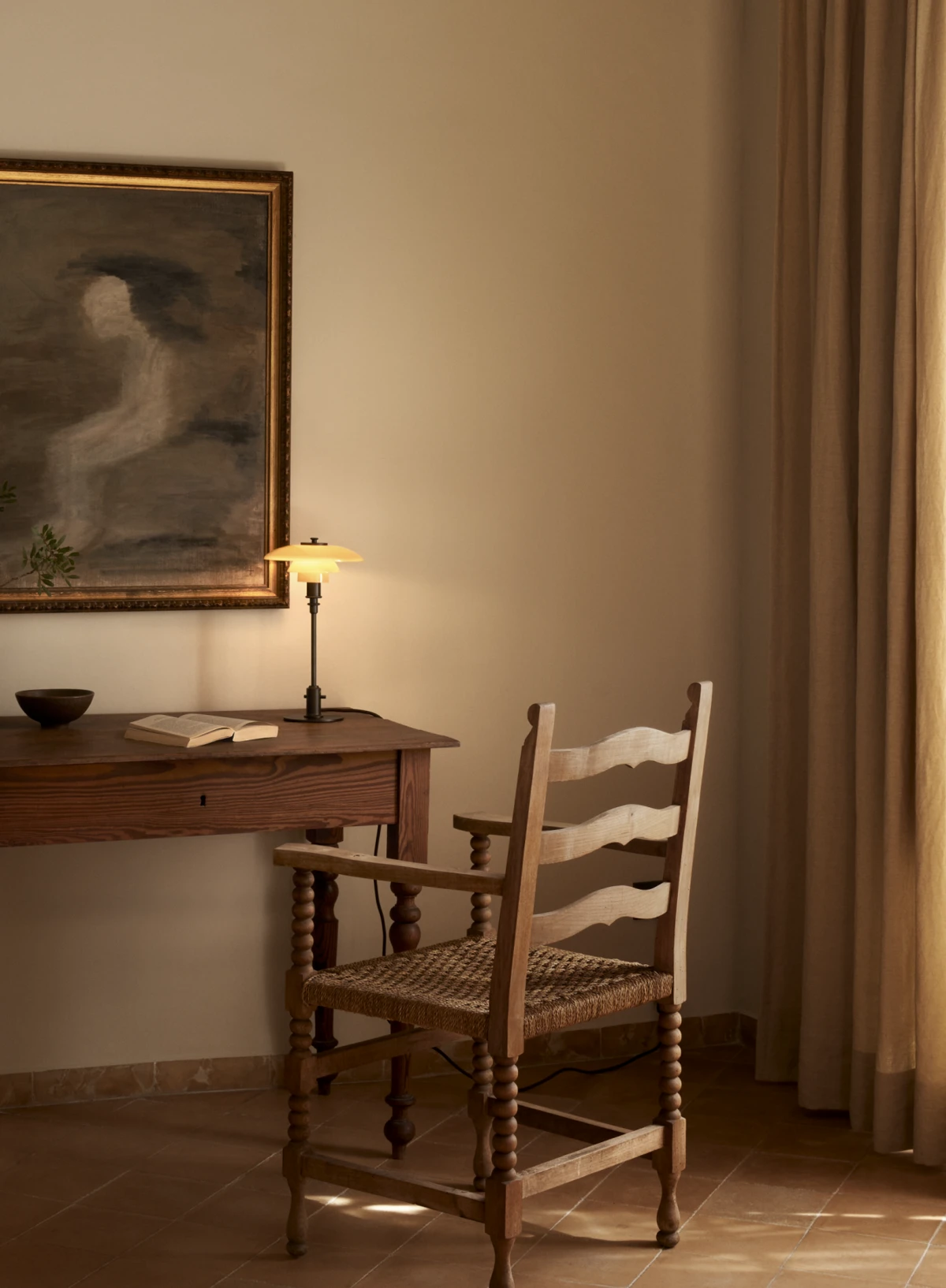 A PH 2/1 Aged Brass Table Lamp casts warm light across an antique wooden desk beside a woven-seat chair. © 2026 Louis Poulsen.