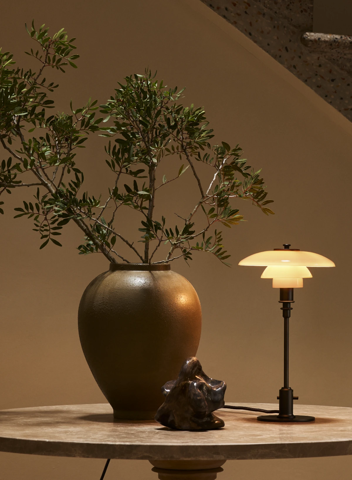 A PH 2/1 Table Lamp with opal glass shades casts a warm glow beside a large vase on a round stone table. © 2026 Louis Poulsen.