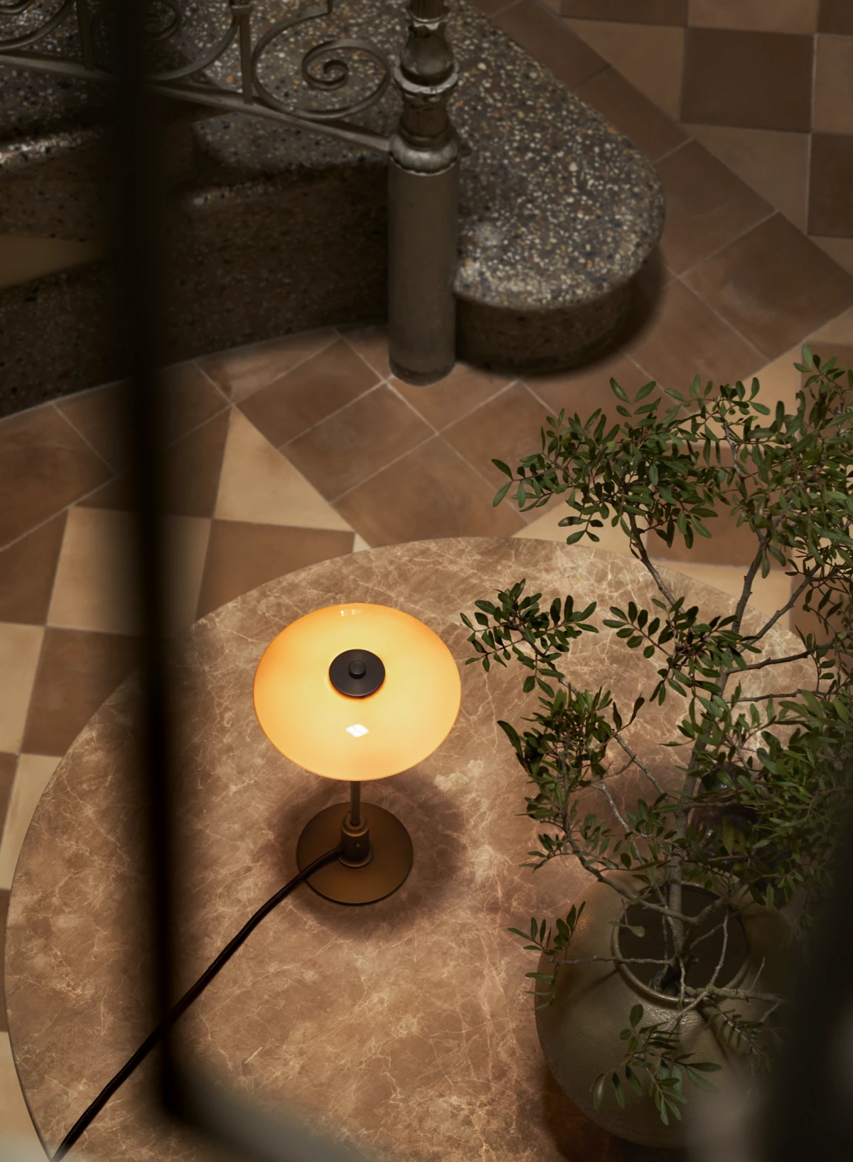 A PH 2/1 Aged Brass Table Lamp casts a warm glow on a marble table beside a leafy plant. © 2026 Louis Poulsen.