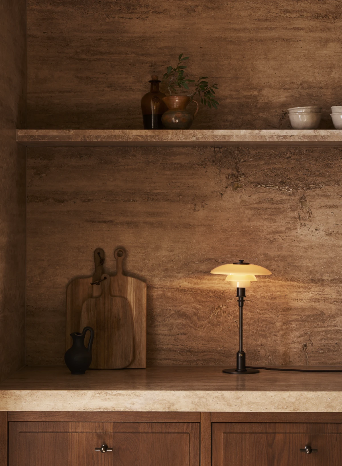 A PH 2/1 Table Lamp casts a warm glow from a dark wood shelf against a brown stone wall. © 2026 Louis Poulsen.