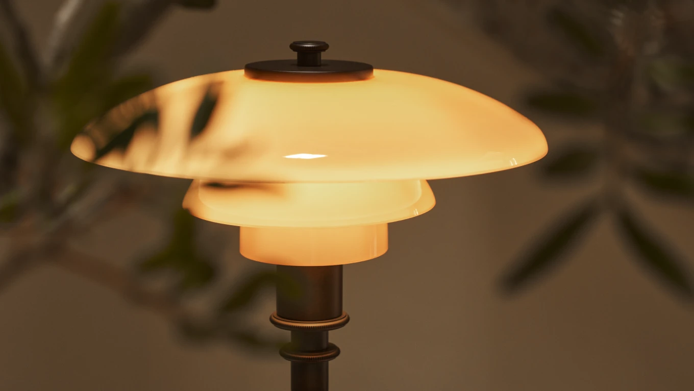 A PH 2/1 Aged Brass Table Lamp glows through amber-coloured glass shades against a dark, leafy background. © 2026 Louis Poulsen.