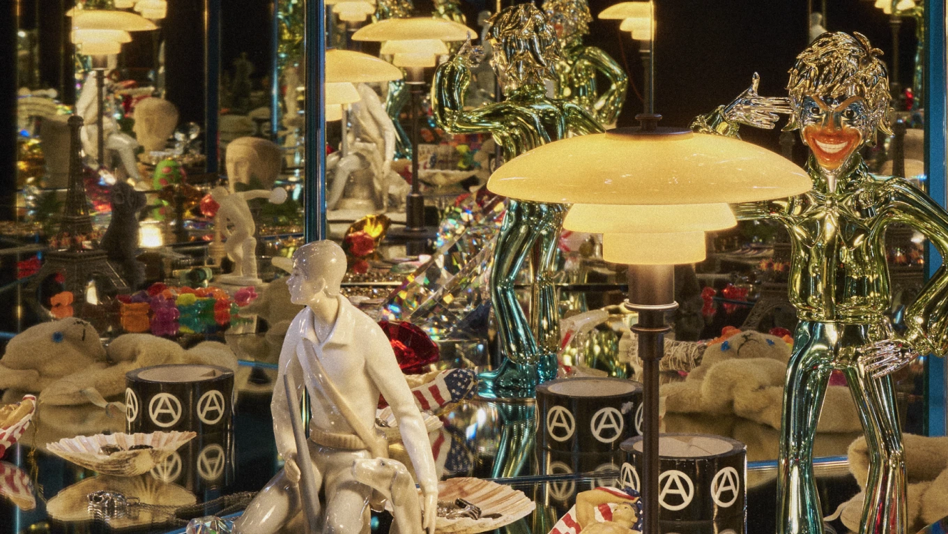 A PH 2/1 Aged Brass Table Lamp casts a warm glow among mirrors, sculptures and colourful objects in a crowded display. © 2026 Louis Poulsen.