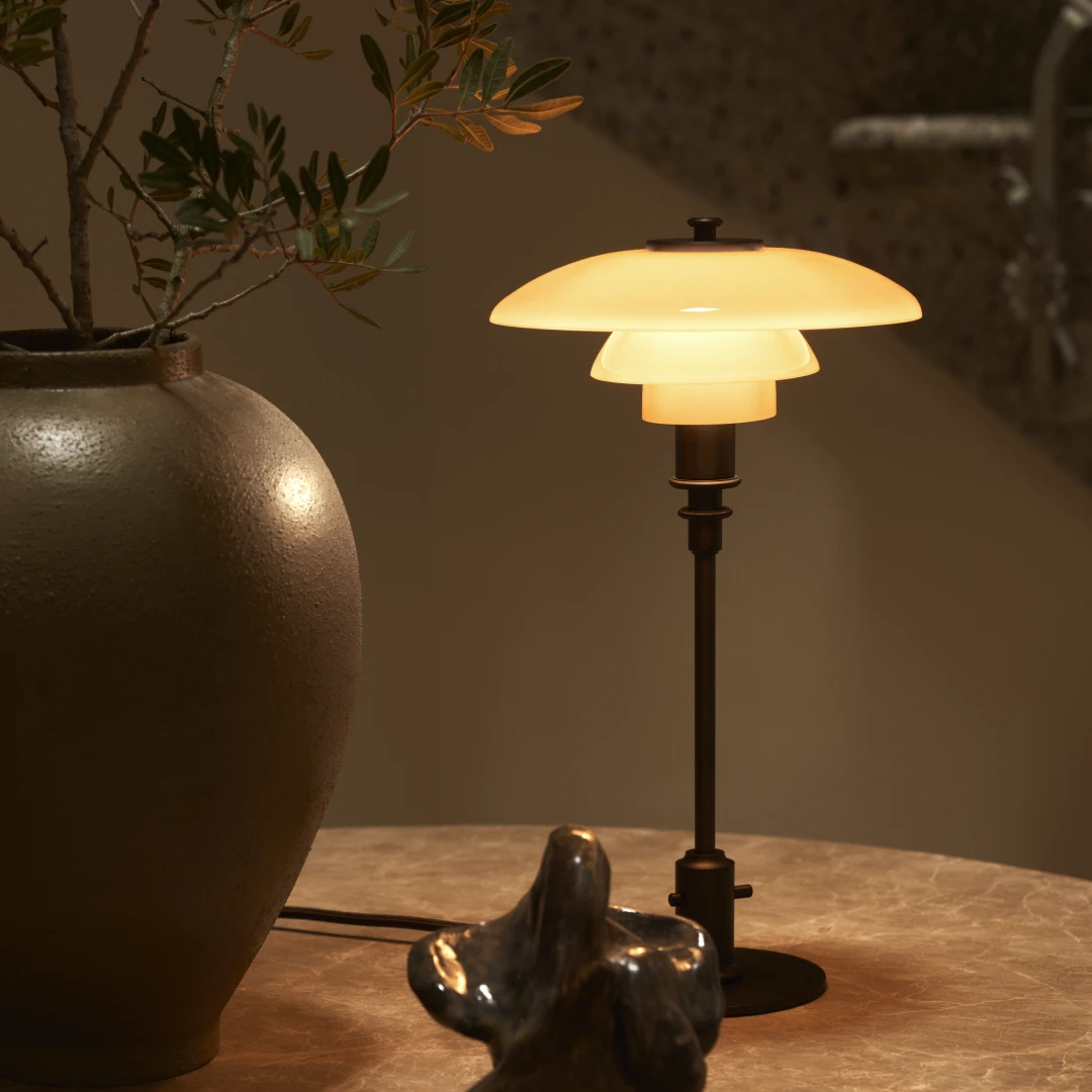 The PH 2/2 Luna Table Lamp glows warmly on a marble table beside a large vase. © 2026 Louis Poulsen.
