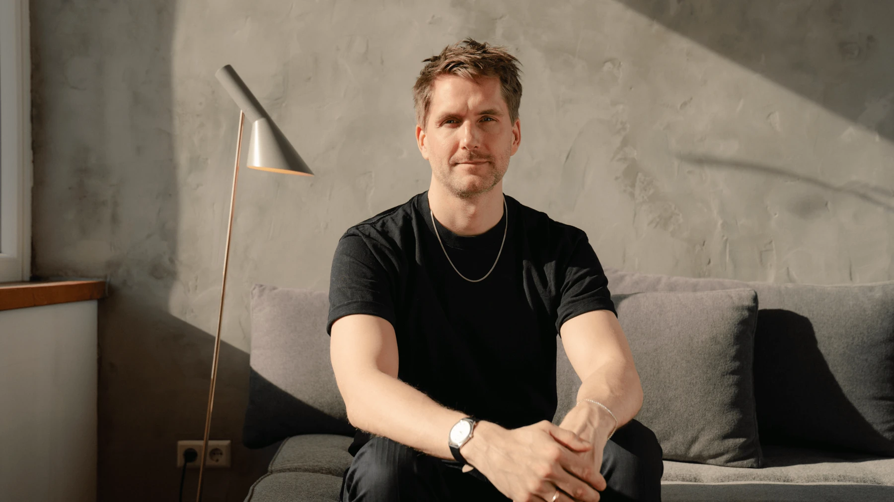 Sigurd Larsen seated beside AJ Floor lamp in natural daylight, © 2025 Louis Poulsen.