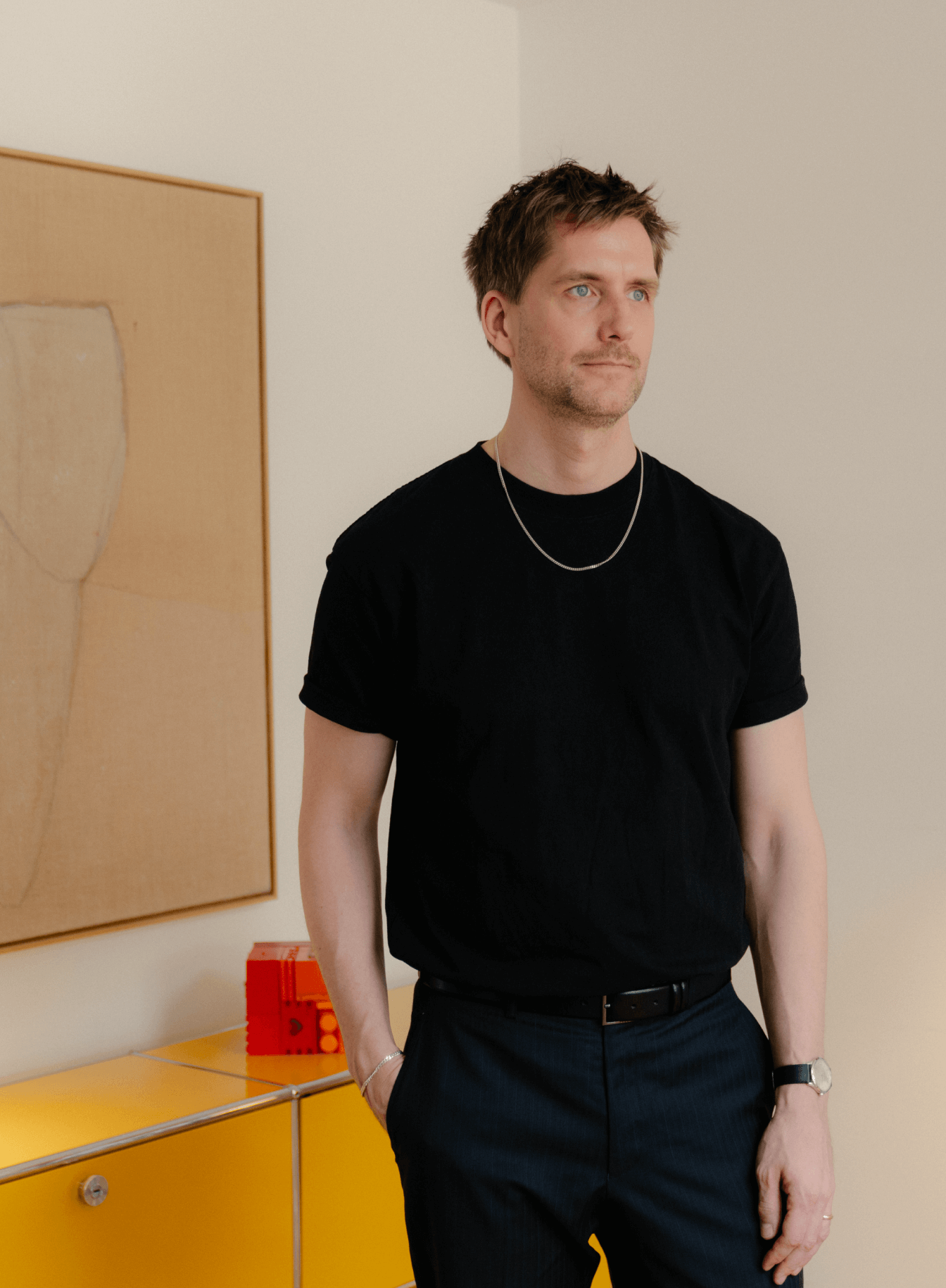 Sigurd Larsen stands beside a yellow cabinet, with framed artwork on the wall behind him. © 2026 Louis Poulsen.