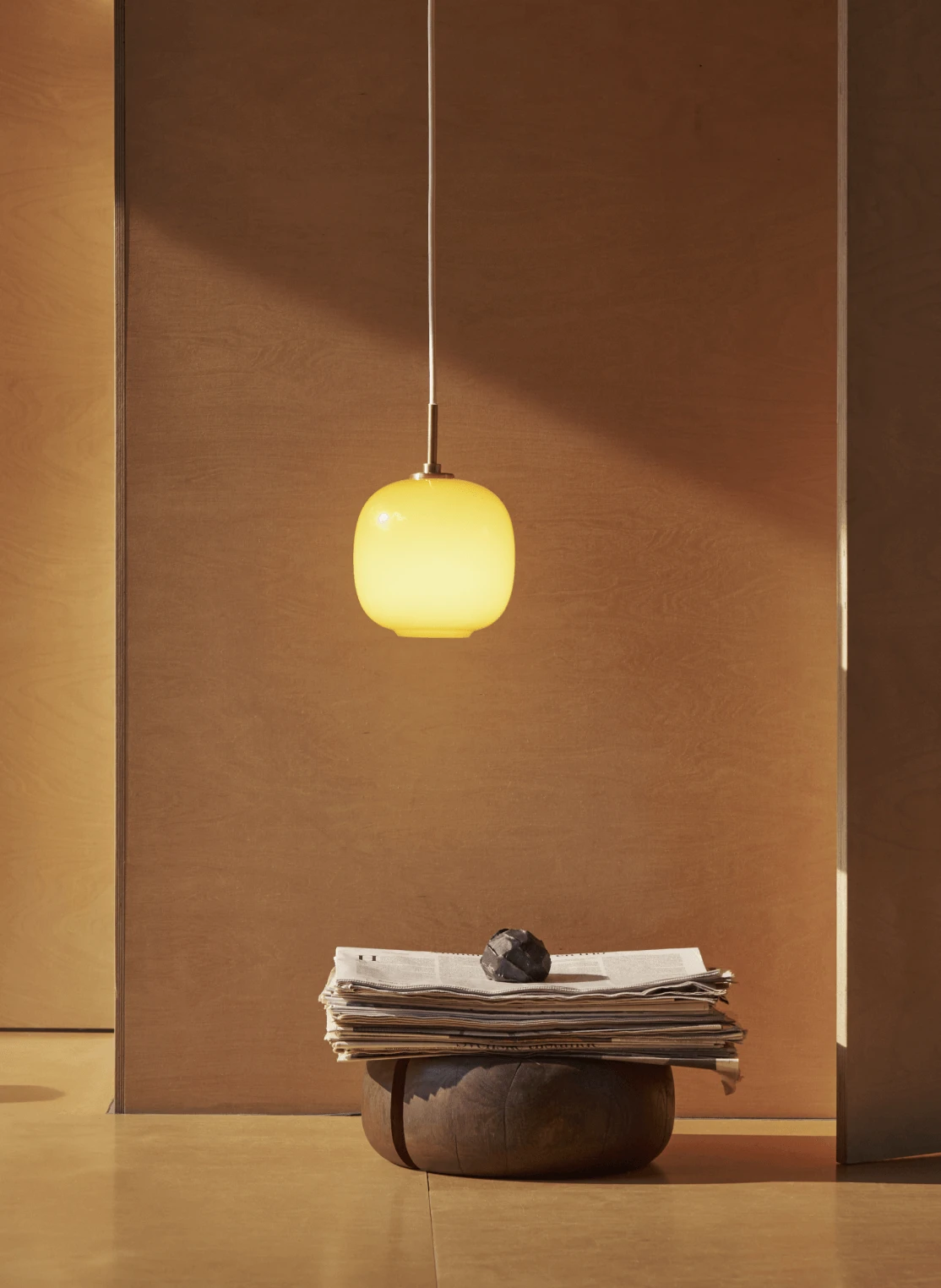 Eine Moser Pendelleuchte aus warm bernsteinfarbenem Glas leuchtet über Zeitungen auf einem niedrigen Tisch vor erdfarbenen Wänden. © 2026 Louis Poulsen.