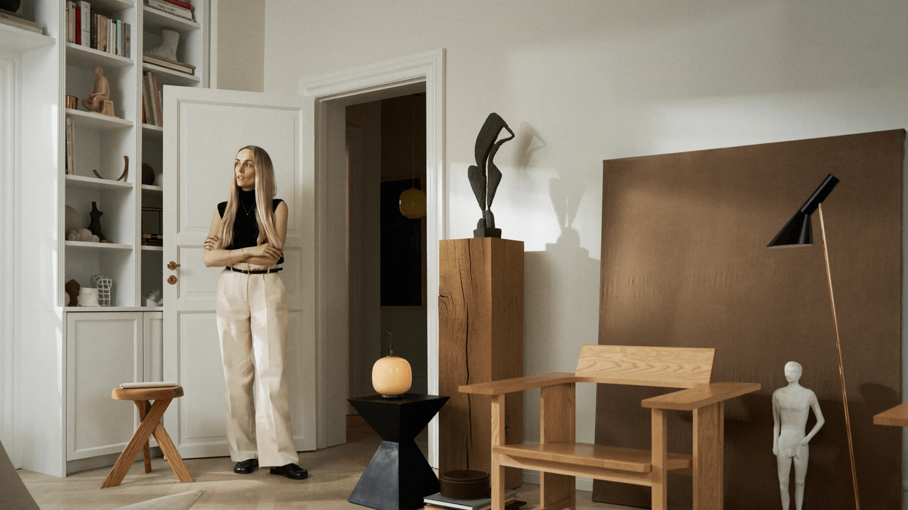 Eine schwarze AJ Floor Lamp steht neben Holzmöbeln; eine VL 45 Radiohus Pendelleuchte leuchtet im Türrahmen. © 2026 Louis Poulsen.