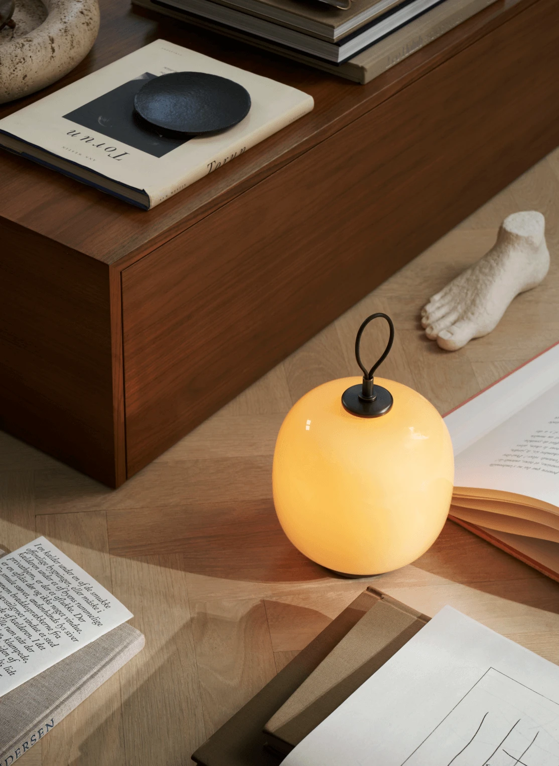 Eine warm leuchtende VL 45 Radiohus Portable Lamp steht zwischen Büchern und skulpturalen Objekten auf einem Holzboden. © 2026 Louis Poulsen.
