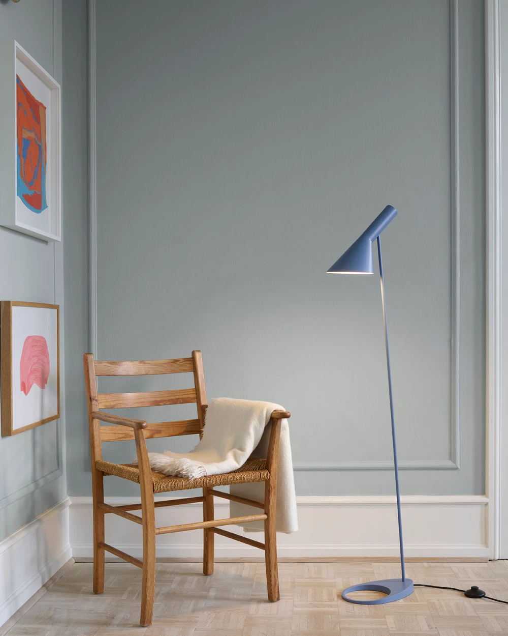 Een blauwe AJ Floor Lamp staat naast een houten stoel met een lichte plaid in een kamer met lichtblauwe muren. © 2026 Louis Poulsen.