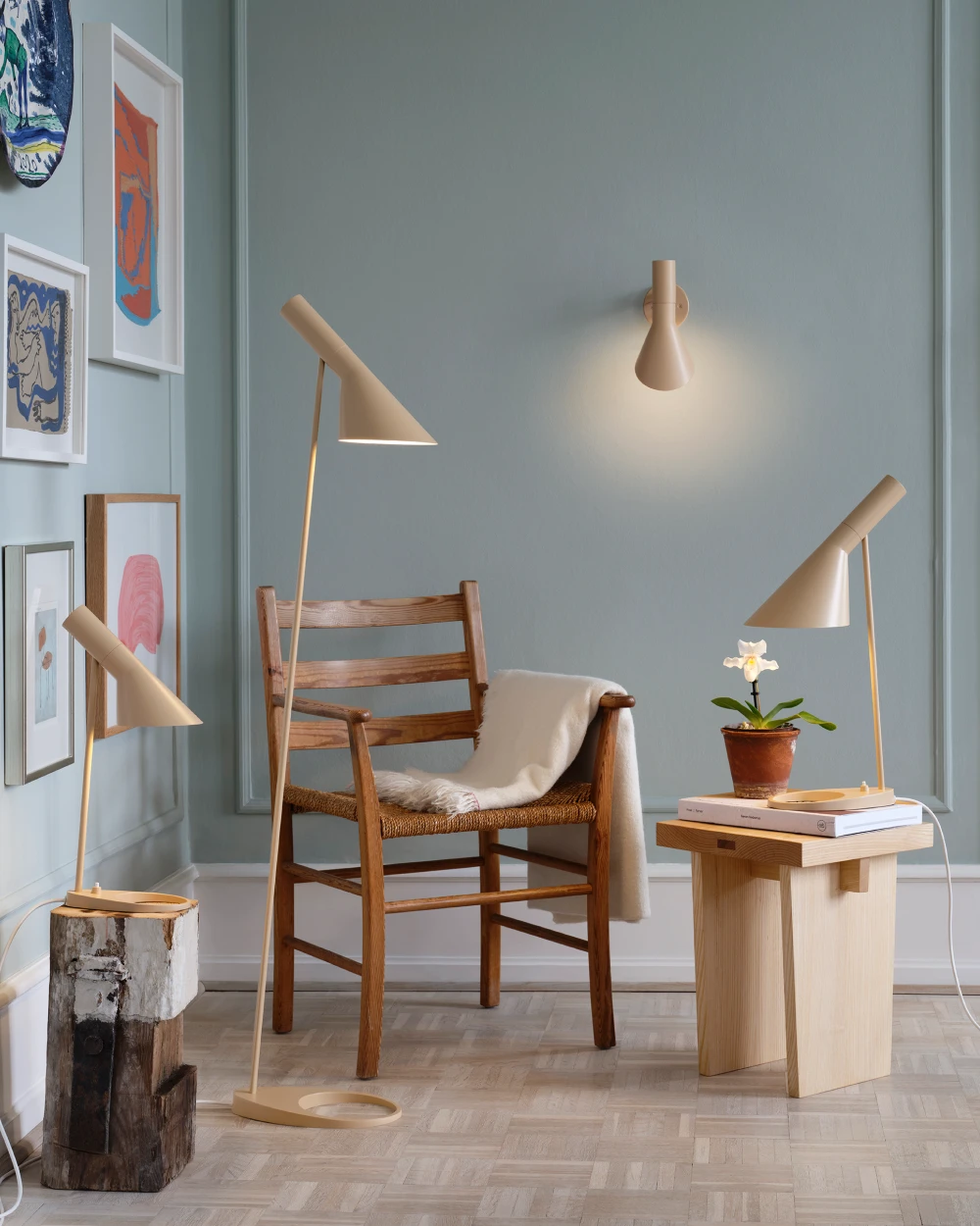 A beige AJ Floor Lamp, two AJ Table Lamp and an AJ Wall Lamp illuminate a living room with wooden furniture and blue-grey walls. © 2026 Louis Poulsen.