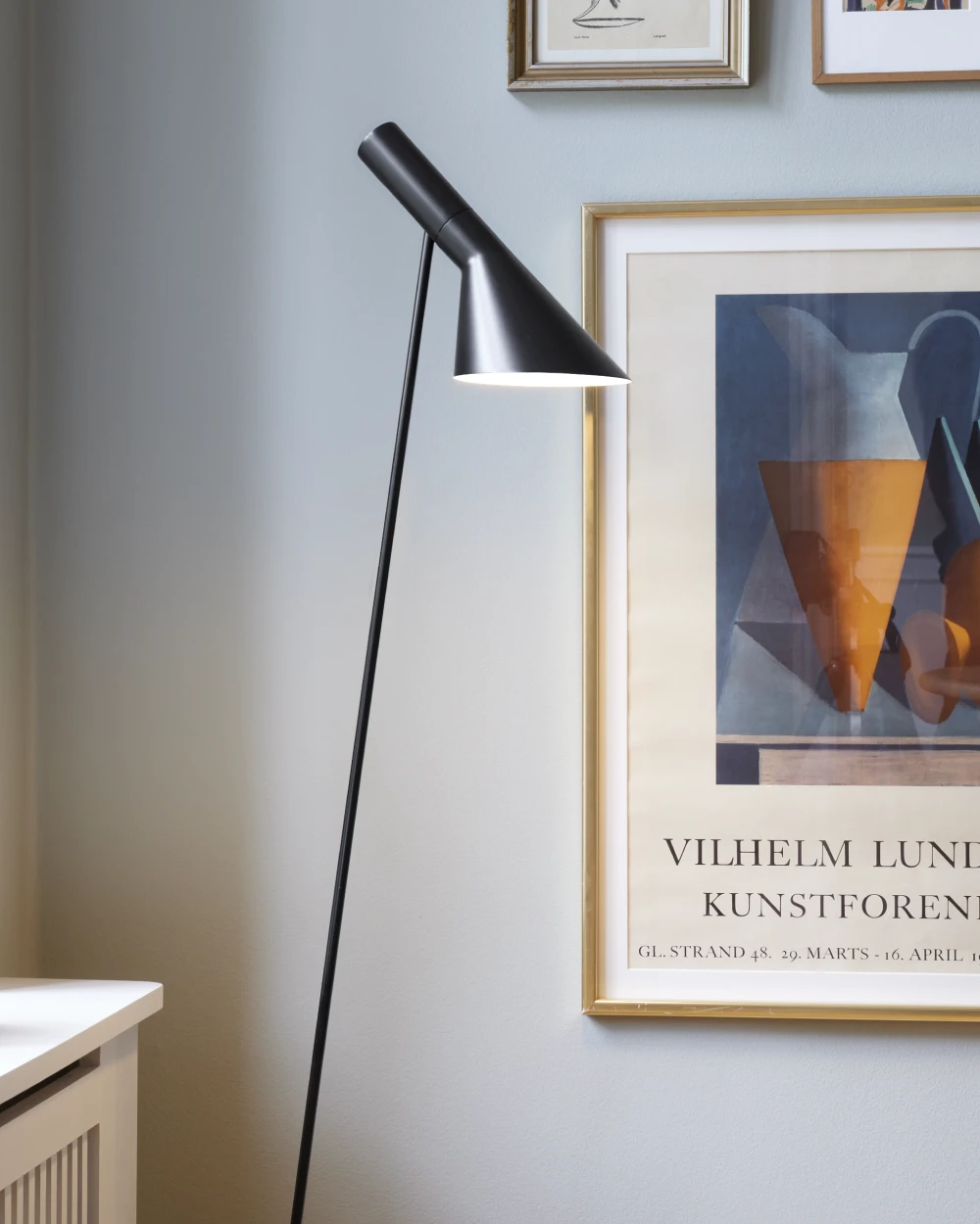 Een zwarte AJ vloerlamp verlicht een lichte muur met ingelijste kunstprints, naast een klein kastje. © 2026 Louis Poulsen.
