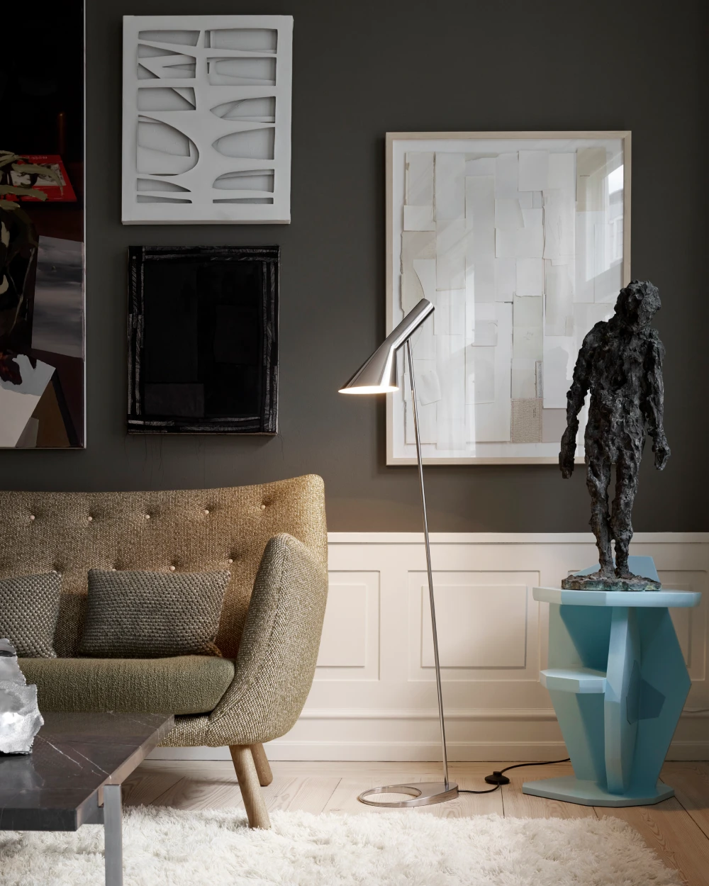 Een chromen AJ vloerlamp verlicht een fauteuil met structuurstof in een woonkamer met ingelijste kunst. © 2026 Louis Poulsen.