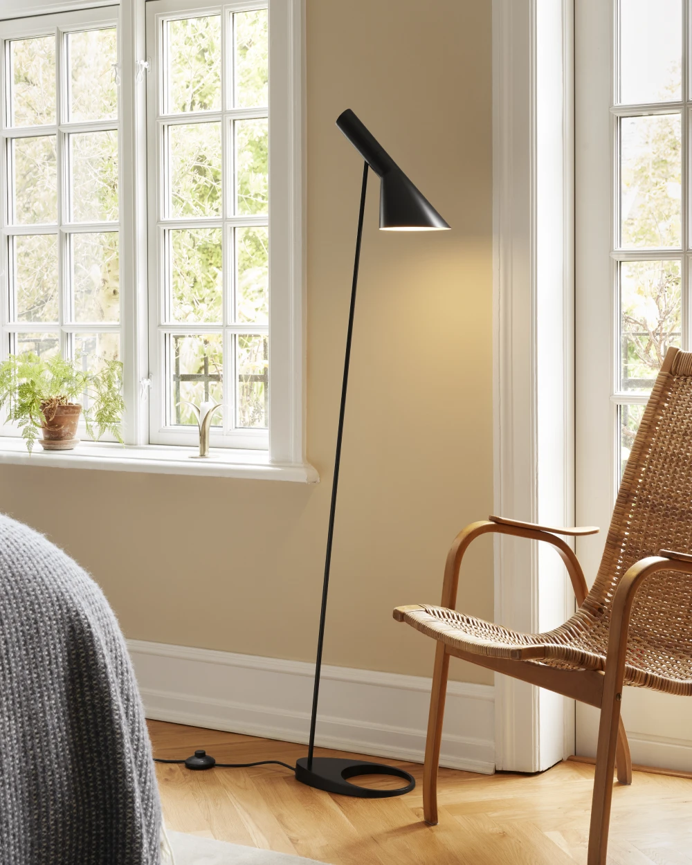 Een zwarte AJ Floor Lamp staat naast een gevlochten fauteuil in een lichte woonkamer met grote ramen. © 2026 Louis Poulsen.