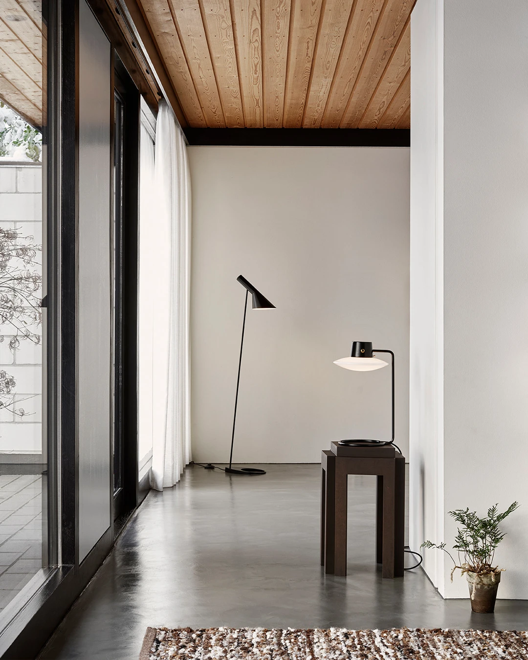 Een zwarte AJ Floor Lamp en een zwarte AJ Oxford Table Lamp staan in een lichte ruimte met een houten plafond en betonnen vloer. © 2026 Louis Poulsen.