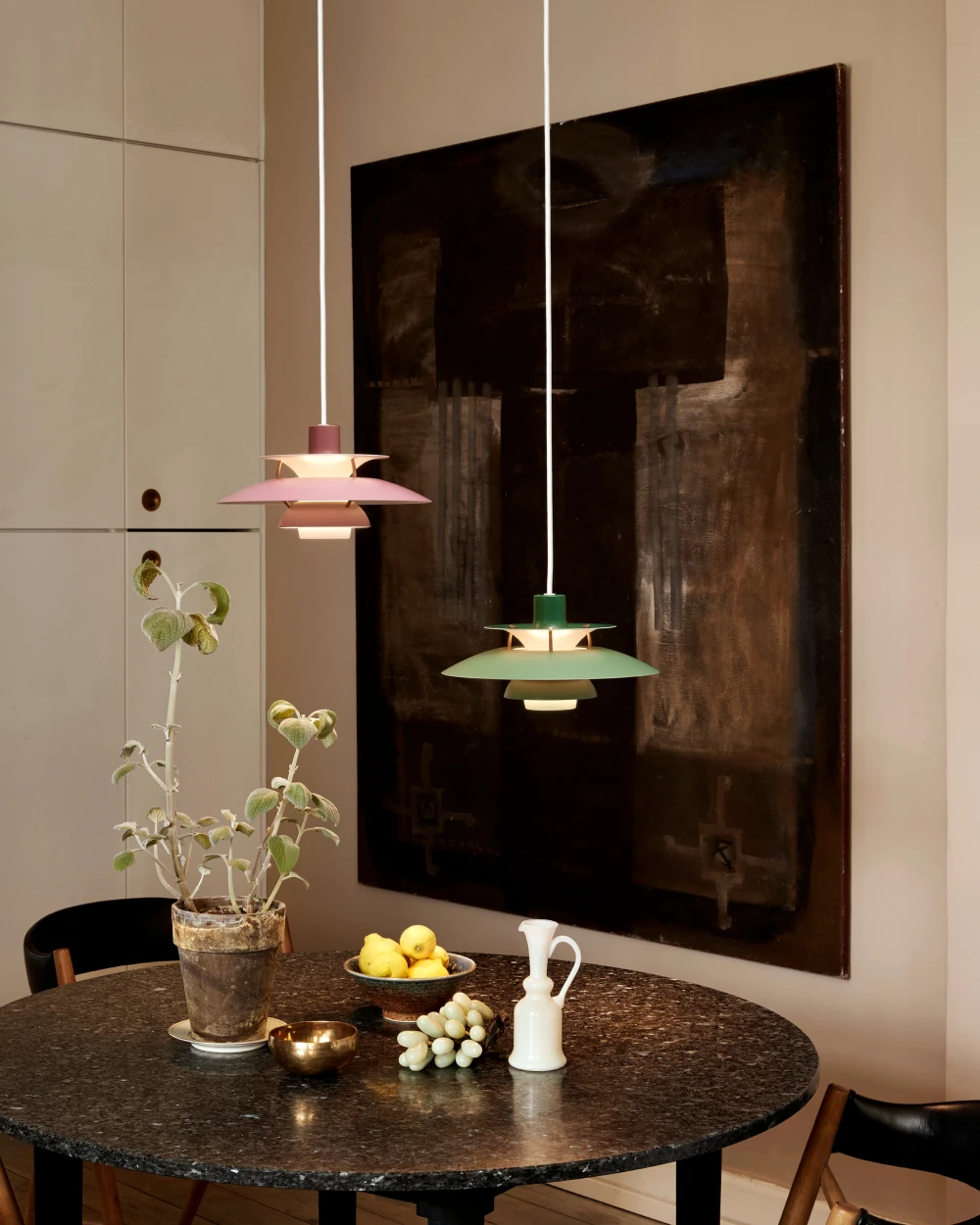 Twee PH 5 hanglampen in roze en groen hangen boven een ronde eettafel met een donker stenen blad. © 2026 Louis Poulsen.