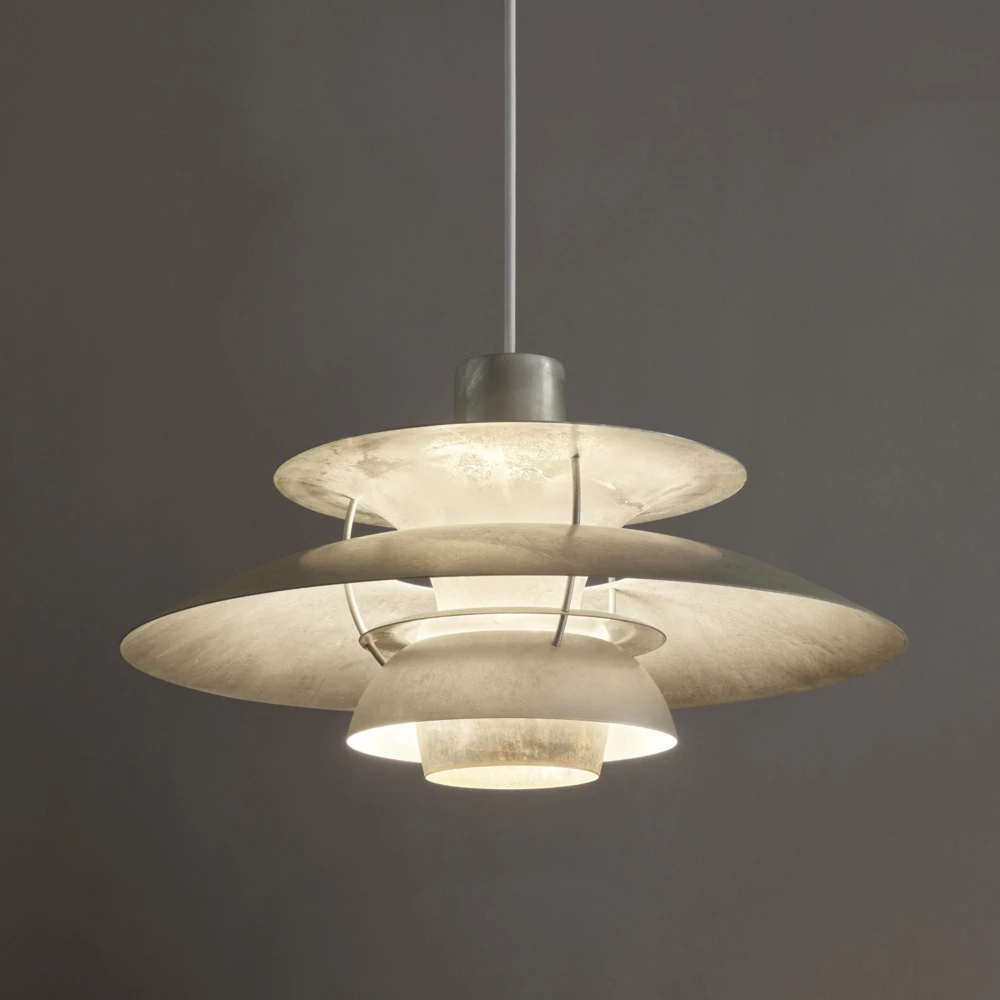 De PH 5 Retake hanglamp met gepatineerde metalen kappen brandt tegen een donkergrijze muur. © 2026 Louis Poulsen.