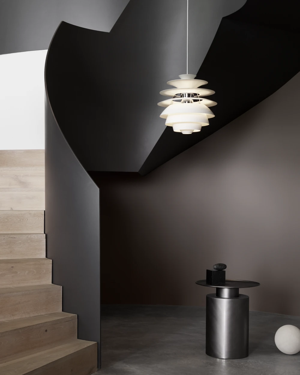 A white PH Snowball pendant hangs in a dark stairwell with pale wooden steps and a metal side table. © 2026 Louis Poulsen.