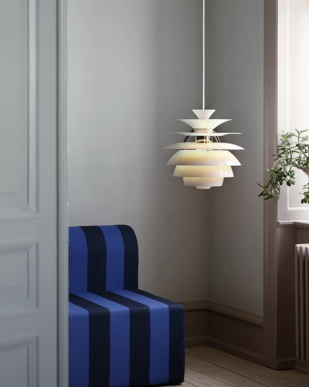 A white PH Snowball pendant glows above a blue-striped chair in a room with grey walls. © 2026 Louis Poulsen.