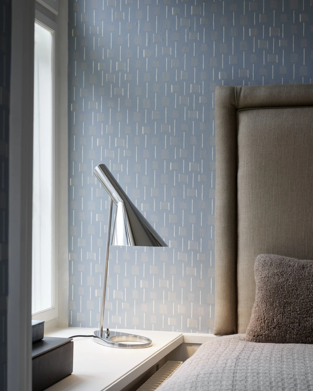 A chrome AJ Table Lamp stands on a bedside table beside an upholstered bed. © 2026 Louis Poulsen.