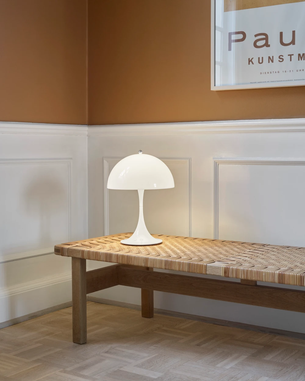 A white Panthella 250 Portable Lamp on a woven wooden table in a room with paneled walls. © 2026 Louis Poulsen.