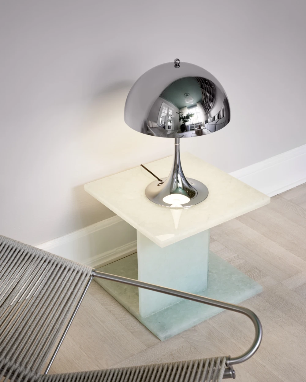 A chrome Panthella 320 Table Lamp sits on a pale stone side table beside a woven chair. © 2026 Louis Poulsen.