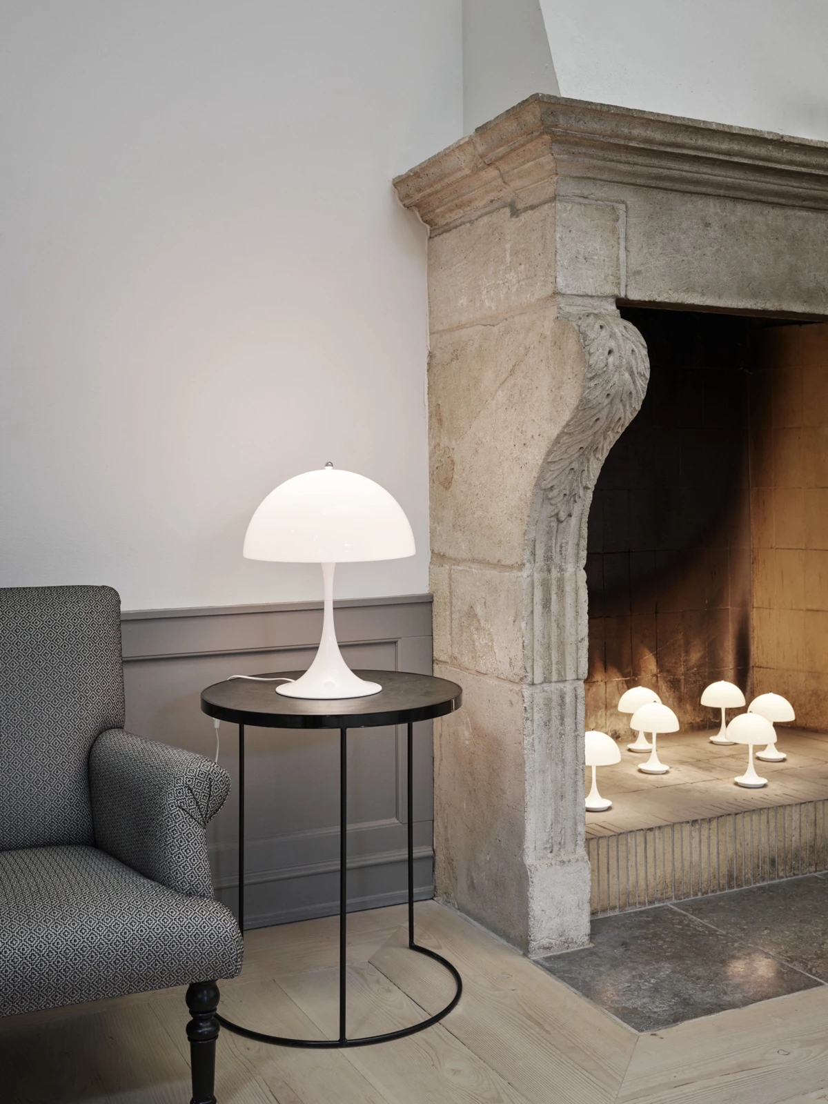 A white Panthella 400 Table Lamp stands beside an armchair and a stone fireplace glowing with smaller lamps. © 2026 Louis Poulsen.