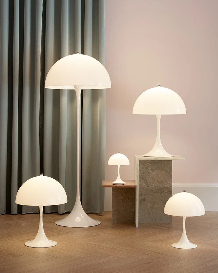 De witte Panthella Floor Lamp en Panthella-tafellampen in verschillende maten staan op een houten vloer bij gordijnen en stenen sokkels. © 2026 Louis Poulsen.