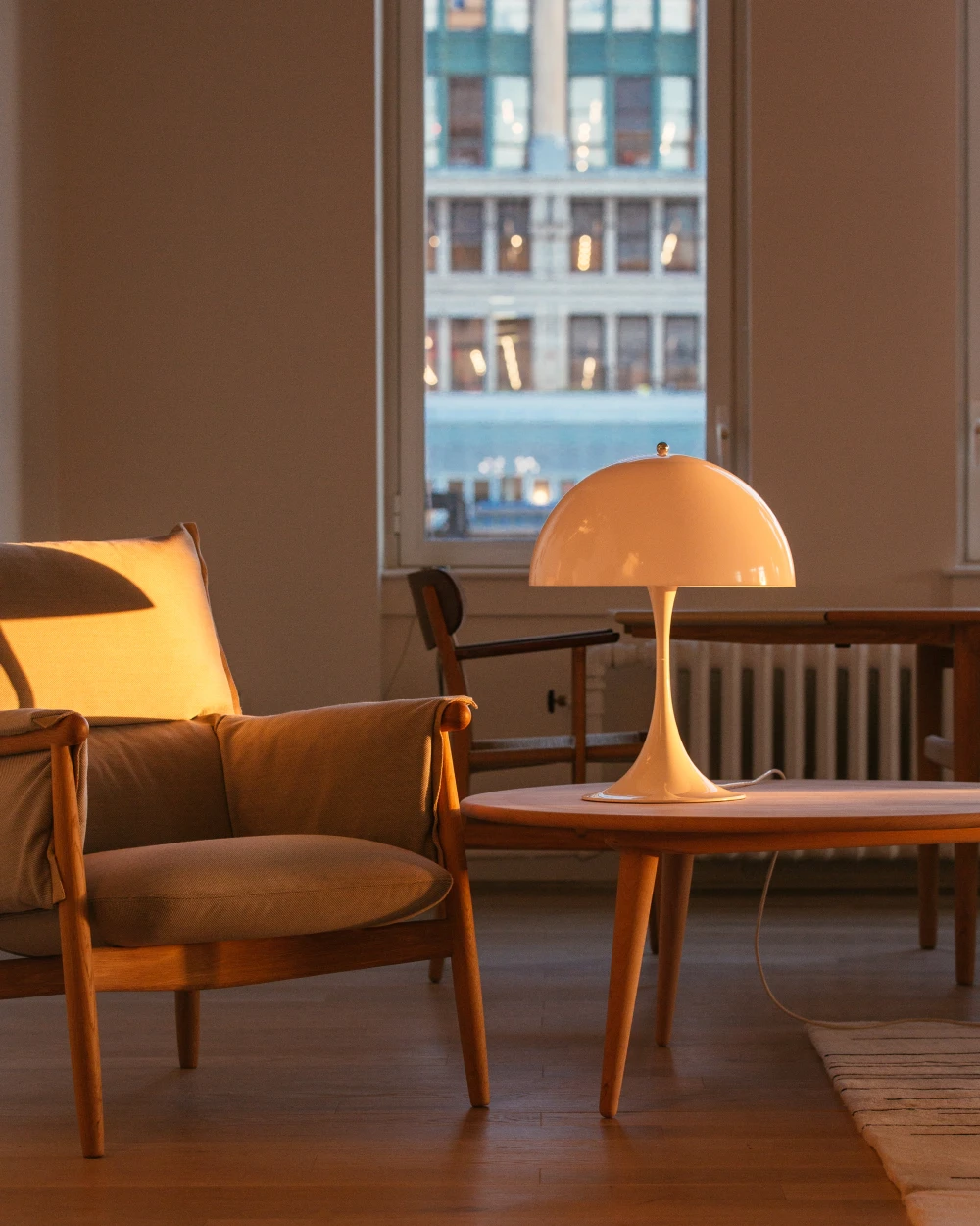 A white Panthella 400 Table Lamp glows on a round wooden table in a living room with an armchair and tall windows. © 2026 Louis Poulsen.